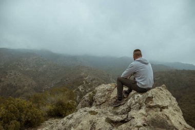 Man from behind in a gray jacket sitting on a rock on top of a mountain in autumn with mist