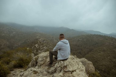 Man from behind in a gray jacket sitting on a rock on top of a mountain in autumn with mist