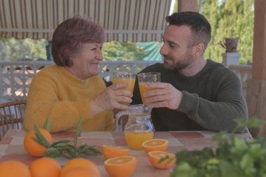 Anne ve oğlu taze sıkılmış portakal suyuyla kahvaltı yapıyor. Doğa ile çevrili bir kır evinde portakallarla çevrili.