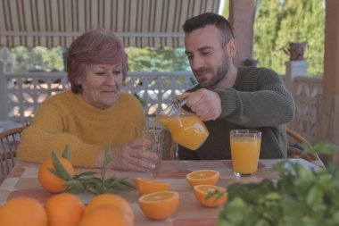 Anne ve oğlu taze sıkılmış portakal suyuyla kahvaltı yapıyor. Doğa ile çevrili bir kır evinde portakallarla çevrili.