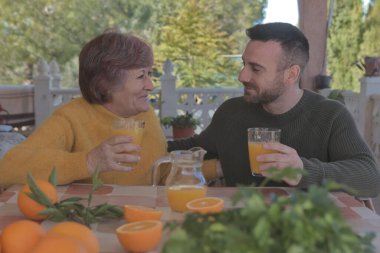 Anne ve oğlu taze sıkılmış portakal suyuyla kahvaltı yapıyor. Doğa ile çevrili bir kır evinde portakallarla çevrili.