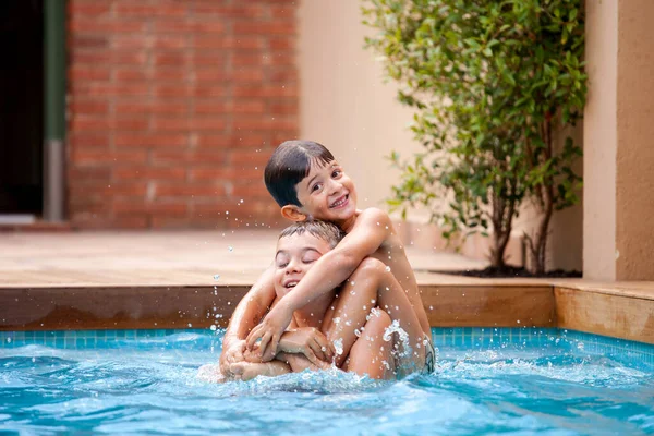 Two siblings playing inside the pool in a sunny summer day. 5-year-old boy on the shoulders of his elder brother in the water. Children relaxing and having fun in vacation. Holidays.