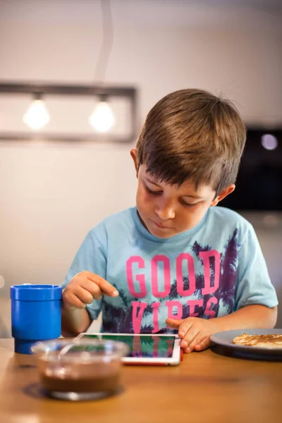 5 years-old kid playing with a digital tablet while having breakfast in a modern kitchen. It\'s daytime in a modern kitchen. Digital education, child development, eating disorders concept.