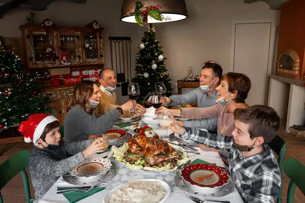 Happy family members wearing surgical masks toasting at the Christmas Thanksgiving table at home in the year of the Covid-19 Pandemic. New rules, new health regulations.