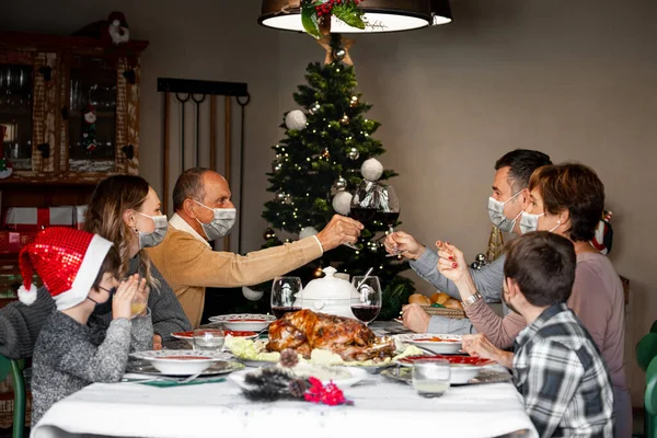 Two family members in surgical masks toast at the Christmas Thanksgiving table at home in the year of the Coronavirus. Happy family members wear surgical masks to comply with new health regulations.