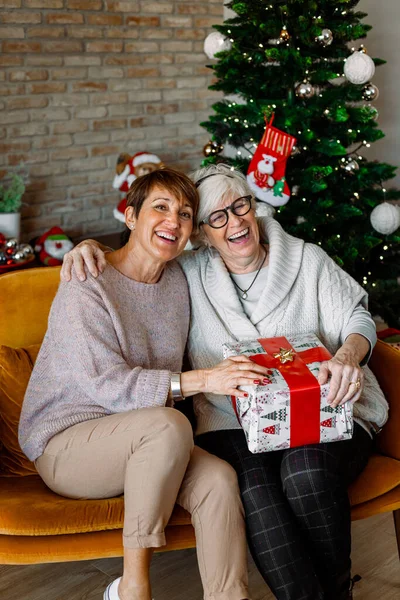 Cheerful excited old ladies sitting on a sofa at the living room, exchanging Christmas gifts. Senior female friends having fun, laughing on Thanksgiving days. Stay active in retirement and friendship.