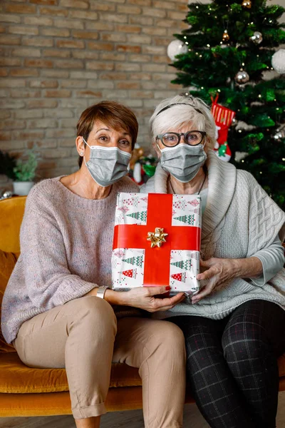 Couple of Elderly female friends, with face mask, exchanging Christmas gifts. Happy senior women with Presents looking at camera. New health regulations during Coronavirus pandemic, lockdown.