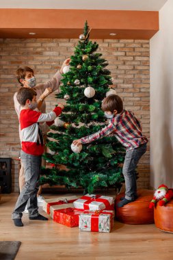 Üç kişilik bir aile, ameliyat maskesi takıyorlar, Noel ağacını evde birlikte süslüyorlar. Büyükanne ve çocuk. Yeni sağlık yönetmelikleri, karantina, Covid salgını, tecrit.