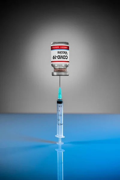 Syringe needle stuck in a vial of red vaccine bottle of COVID-19. Syringe standing vertical on a blue reflective surface. Studio background. Cure for COVID-19 pandemic virus, medical crisis.