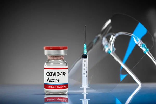 Covid-19 vaccine vial, syringe and face shield protector on a reflective blue surface. Protection medical equipment for vaccination. Still life shot. Cure for Pandemic COVID-19 virus, medical crisis.