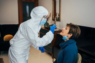 Health Professional in PPE, face mask and shield, introducing a nasal swab to a senior female patient on a home visit. Rapid Antigen Test to analyze nasal sampling for Covid-19, Coronavirus Pandemic. clipart