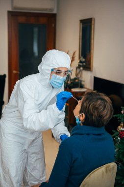 Health Professional in PPE suit, mask and face shield introducing a nasal swab to a senior patient at her house. Rapid Antigen Test kit to analyze nasal culture sampling while coronavirus Pandemic. clipart