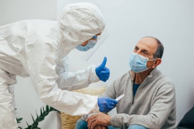 Health professional in a PPE suit, mask, face shield showing the patient the negative result for Covid-19. Antigen test to an elderly patient during a home visit. Coronavirus Pandemic. clipart