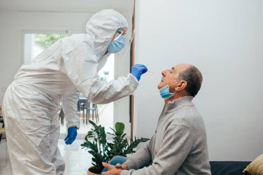Health Professional in PPE suit and face shield introducing a nasal swab to a senior adult patient at his house. Rapid Antigen Test kit to analyze nasal culture sampling while coronavirus Pandemic. clipart