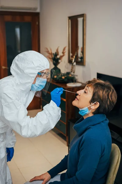 Health Professional in PPE, face mask and shield, introducing a nasal swab to a senior female patient on a home visit. Rapid Antigen Test to analyze nasal sampling for Covid-19, Coronavirus Pandemic.