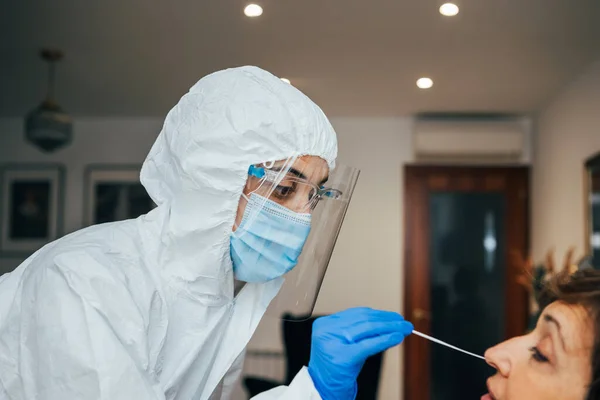 Close up of female health Professional in PPE, face mask and shield, introducing a nasal swab to a senior female patient on a home visit. Rapid Antigen Test kit to analyze Covid-19.