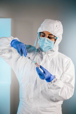 Covid-19 aşı şişesine ve araştırma laboratuarında şırıngaya bakan kadın bilim adamı. PPE giyiyor, yüz koruyucusu ve tıbbi eldiven takıyor, şişeden ilaç alıyor..