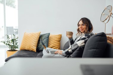 Rahat genç bir kadın rahat bir kanepede oturuyor, elinde bir fincan kahveyle, dairede dizüstü bilgisayarla çalışıyor. Freelancer ya da kameraya bakan öğrenci. Coronavirus salgını sırasında evden çalışmak.