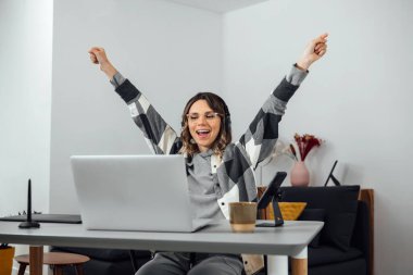 Heyecanlı bir kadın, kolları açık, ev ofisinde oturuyor. İşkadını Serbest Çalışan Yükseltilmiş Ellerle Başarı Kutlaması. Sevinçten havalara uçuyor, laptopta iyi haberler alıyor, zafer, piyango kazanıyor..