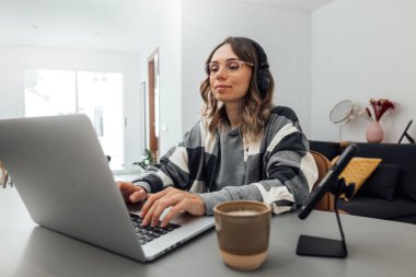 Kafkasyalı genç bir kadın, kulaklık takıyor ve ev kıyafetleri giyiyor, İçişleri Bakanlığı 'ndaki bir dizüstü bilgisayarın klavyesinde daktilo yazıyor. Kız öğrenci, serbest çalışan, çalışıyor, online iş yapıyor. Coronavirus kilitlenmesi.