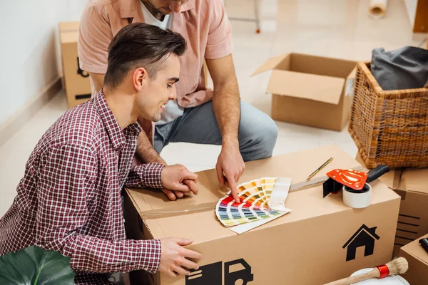 Close up of Young gay couple holding hands, sitting on the floor of a apartment they moved in, and choosing a color from a color pattern. Moving into a new home concept, renovation, designing..