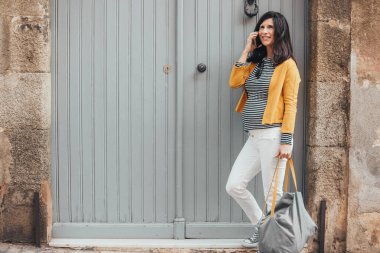 Young pregnant fashionable woman in stripes shirt, yellow jacket and bag, standing in front a door of an old town street, talking on the phone. Smiling brunet woman using mobile. Copy-space.