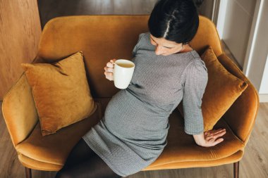Top view of a Stylish caucasian brunet pregnant woman in black and white dress, sitting in a mustard velvet sofa, drinking a cup of tea or coffee. Expecting mother resting, taking a break from work.