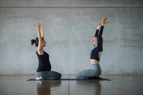 Smiling young sportive women training pilates together, sitting on their knees on a mat on the floor, with arms up, doing a yoga posture. Pregnant woman working out with personal yoga trainer.