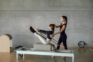 Koruyucu maskeli bir kadın Reformist makinesinde kişisel pilates dersi alıyor, büyük bir spor salonunda, geniş endüstriyel spor salonunda. Sağlıklı fitness yaşam tarzı.