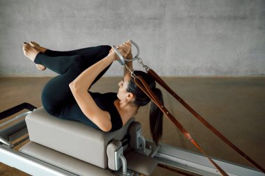 Esmer, sportif kadın, deri bantları ve ipleri pilates makinesinden çekiyor. Profesyonel bir koçun spor salonunda, spor salonunda vücut ve kol egzersizi yapması..