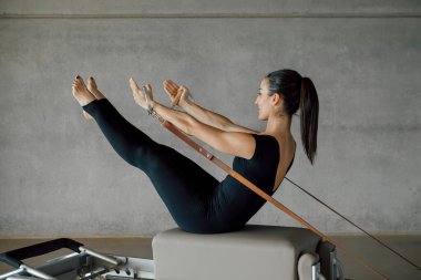 Bir pilates eğitmeninin güçlü vücudunun, bir Reformist makinesinin yatağında uzanmış koli bandını elleriyle çekerken, büyük bir endüstriyel fitness salonunda antrenman yaparken. Tam uzunluk.