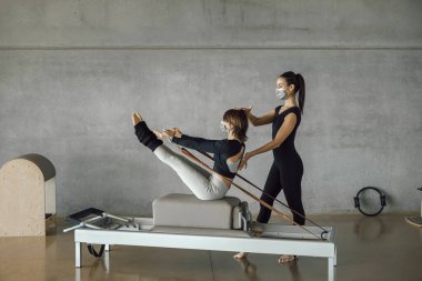 Koruyucu maskeli bir kadın Reformist makinesinde kişisel pilates dersi alıyor, büyük bir spor salonunda, geniş endüstriyel spor salonunda. Sağlıklı fitness yaşam tarzı.