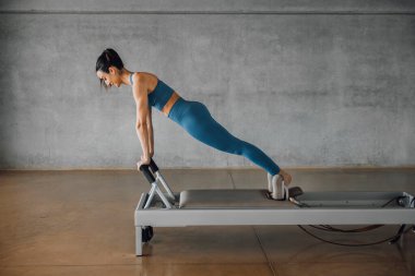 Spor giyimli kararlı genç bir kadının spor salonundaki Pilates Reformer Machine Yatağı 'nda tahta pozisyonu. Büyük endüstriyel çatı katında eğitim..