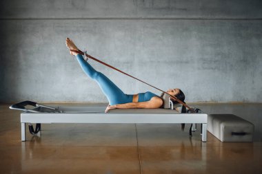 Pilates Reform Makinesi 'nin yatağında uzanmış kadın pilates eğitmeni ayaklarıyla, bacaklarıyla bandı çekerken, büyük bir endüstriyel spor salonunda antrenman yaparken yan görünümü. Tam uzunluk.