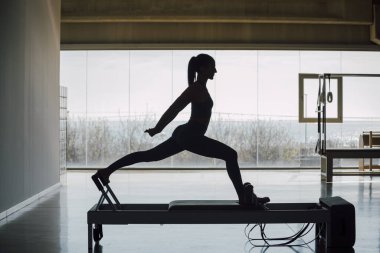 Reformer Yatak Makinesi 'nde pilates egzersizi yapan formda bir bayan koç. İnce bir kadın, büyük pencereli ve sokak manzaralı büyük bir spor salonunda bacaklarını esnetiyor..