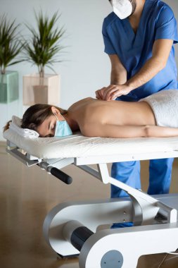 Erkek fizyoterapist, ameliyat maskesi takan ve sırt üstü sedyeye uzanmış genç bir kadına masaj yapıyor. Sağlık ve sağlık merkezinde sırt ağrısı ve boyun ağrısı için elle tedavi uyguluyor..