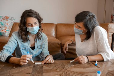 Two young women friends in protective face mask sitting together at home, realizing a self Antigen test for Coronavirus COVID-19. Woman manipulating the nasal sample tube and the device.