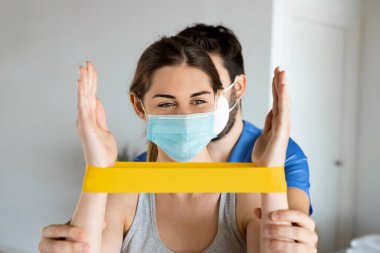 Fizyoterapist ya da doktorla bireysel egzersizler sırasında sarı elastik bantla gerinen gülümseyen genç bir kadının portresi. Tedavi kliniğinde ya da sağlık merkezinde rehabilitasyon.
