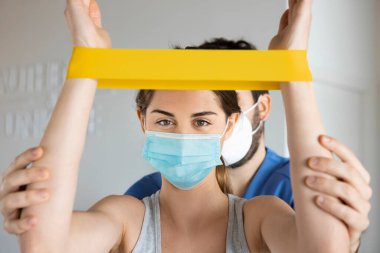 Lastik bant kullanarak rehabilitasyon yapan genç bir kadın, klinikte ya da hastanede fizyoterapistle yüz maskesi takıyor. Covid-19 salgını sırasında fizik tedavi.