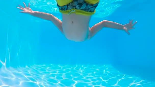 スローモーション 低角度 黄色の水着の少年は バブルと太陽の光で 暑い晴れた日にリフレッシュするために青い澄んだプールの水にダイビングします 夏休みを楽しむ子供 — ストック動画