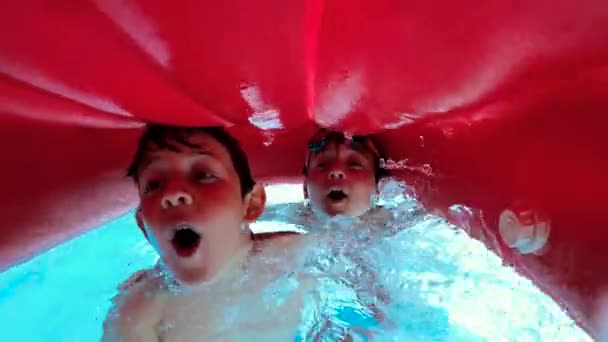 SLOW MOTION, UNDERWATER, PORTRAIT: Zwei glückliche Jungs, die im Pool schwimmen und lächelnd winken. Kinder atmen tief durch, um in den Sommerferien ins kristallklare Wasser zu gelangen. Kindheit, Freundschaftskonzept