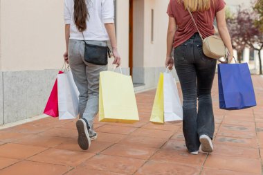 İki genç kadın ellerinde rengarenk alışveriş torbalarıyla yaya kaldırımında yürürken arkadan görülüyor. Kentsel ortam, günlük kıyafetler ve rahat bir atmosfer. Yatay, arka plan.