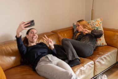Two friends on a leather sofa posing for a selfie. One smiling with tongue out, the other making a peace gesture. Concept of fun, bonding, youth lifestyle and social media. Horizontal, close-up view.