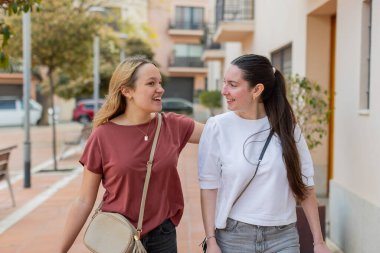 İki bayan arkadaş yan yana yürüyorlar, gülümsüyorlar ve şehir merkezinde dostça sohbet ediyorlar. Yatay, göz hizasında görünüm.