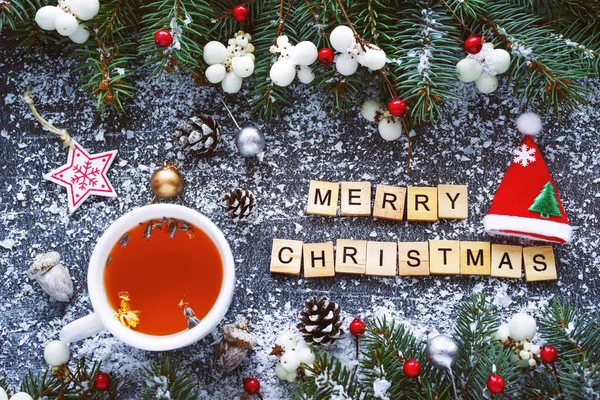 Cup of tea and text from wooden letters Merry Christmas on a textured surface surrounded by Christmas decor and fir branches, top view. Christmas background.