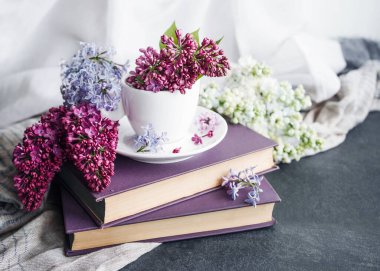 Bahar şenliği arka planında beyaz bir fincanda leylak çiçekleri, bir yığın kitabın üzerinde narin örtülü kumaş, yakın plan, yumuşak odak