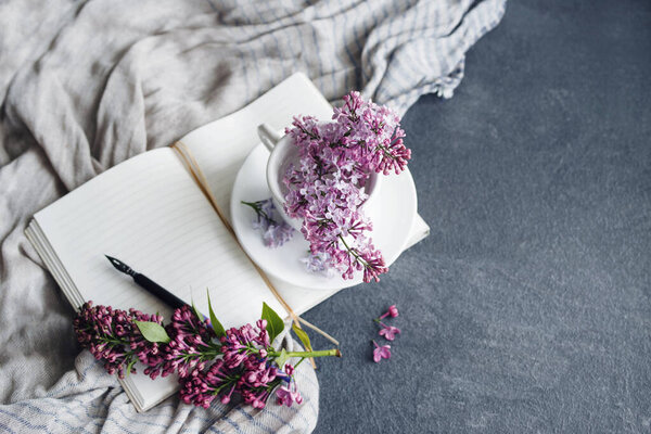 Весенний романтический фон с сиреневыми цветами в белой чашке, с открытой белой тетрадкой и ручкой для рисования на фоне нежной драпированной ткани на сером столе, вид сверху, пространство для копирования