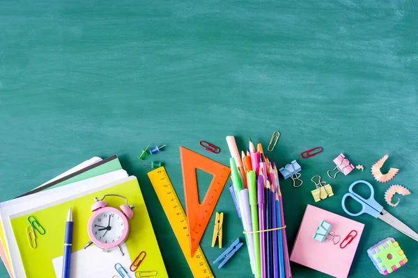 Back to school background. School supplies, pens, pencils, notebooks and an alarm clock on a  against the background of a green school board, copy space, top view.
