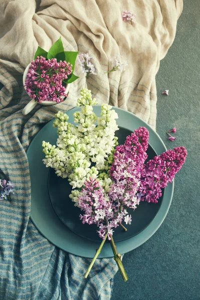 Yuvarlak gri tabaklar üzerinde beyaz ve pembe leylak demeti ve koyu bir yüzey üzerinde çizgili pamuklu şal, üst manzara, tonlu