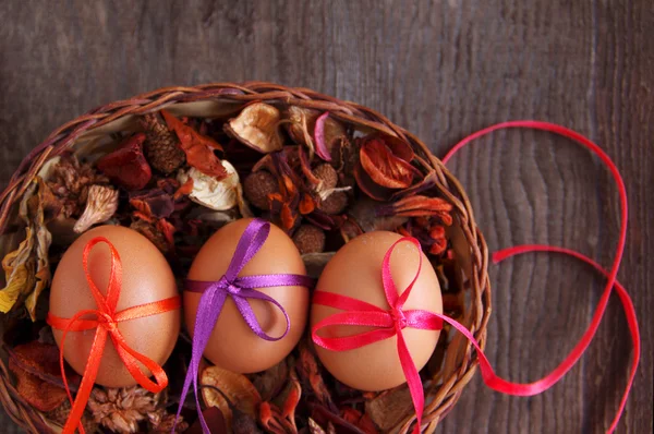 Três ovos de Páscoa decoram-se brilhantes com fitas a cores em uma cesta com flores secas . — Fotografia de Stock